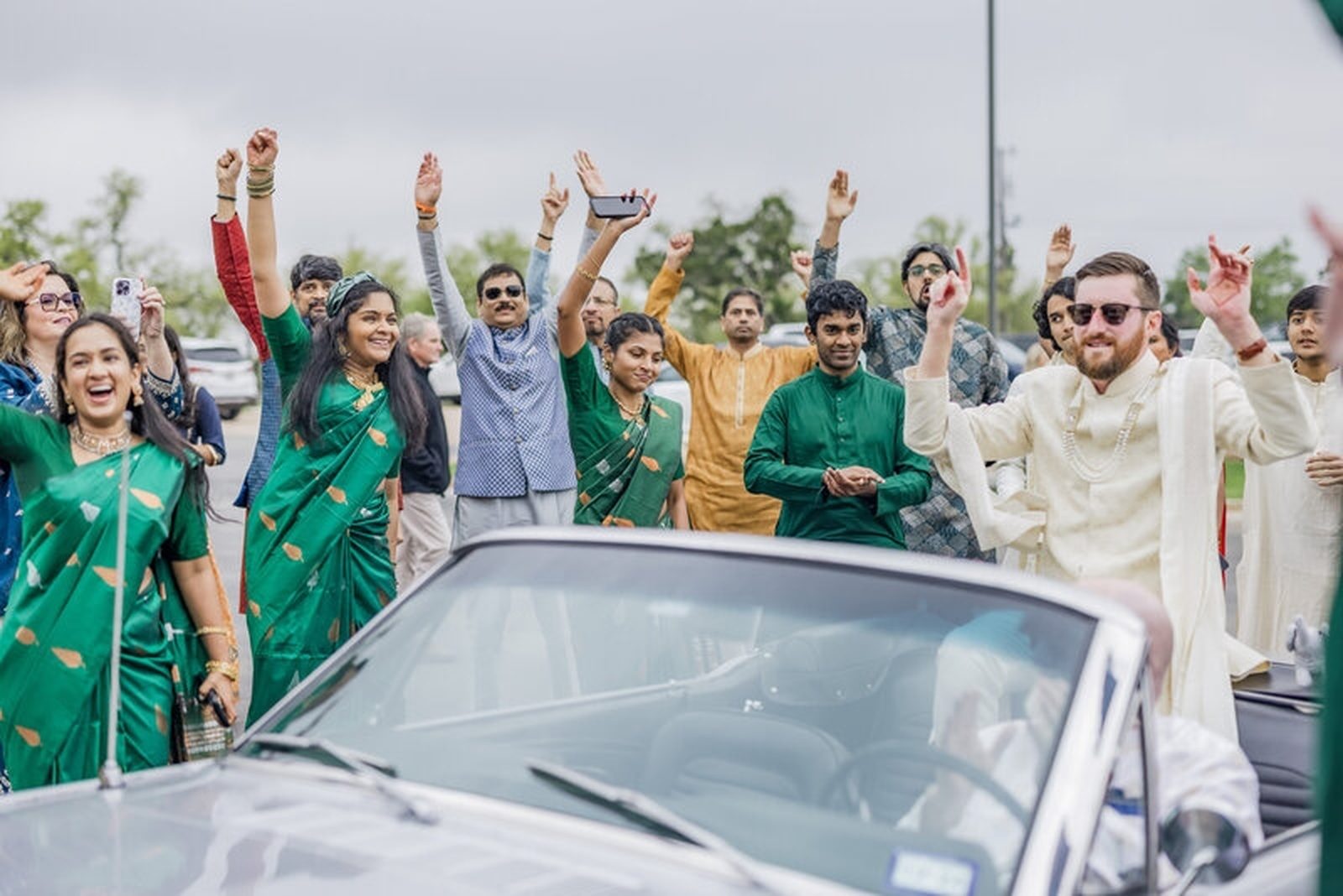 Guests cheering with raised hands around the groom in a vintage convertible during the baraat