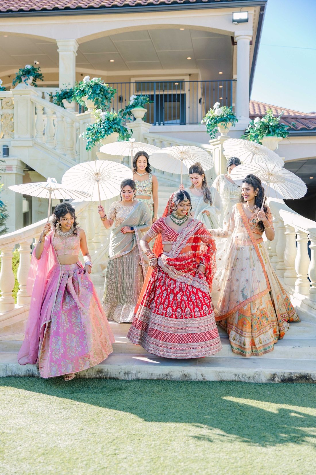 Bride in red descending an outdoor staircase surrounded by her bridal party holding parasols