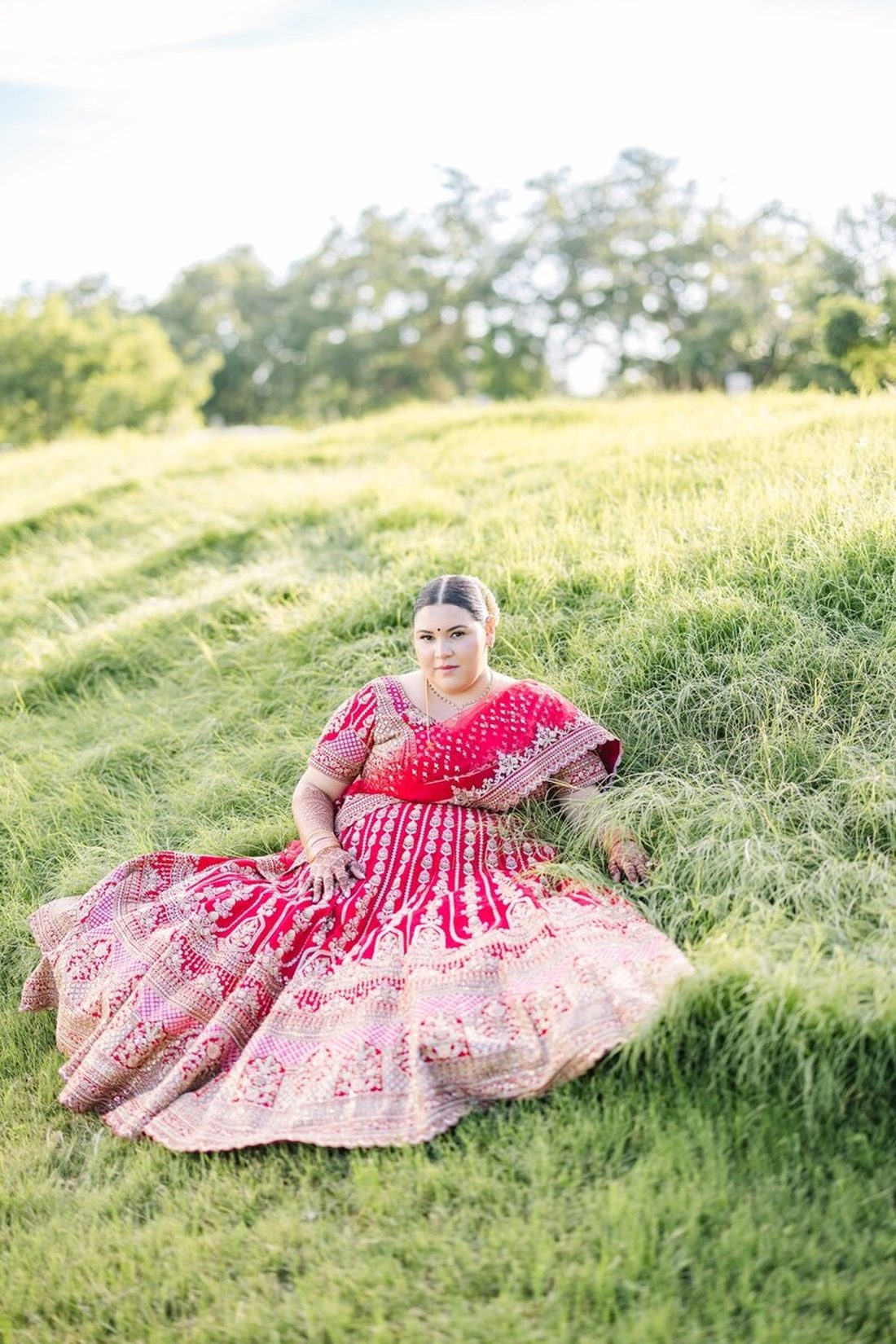 Bride in a crimson lehenga seated in tall green grass at golden hour