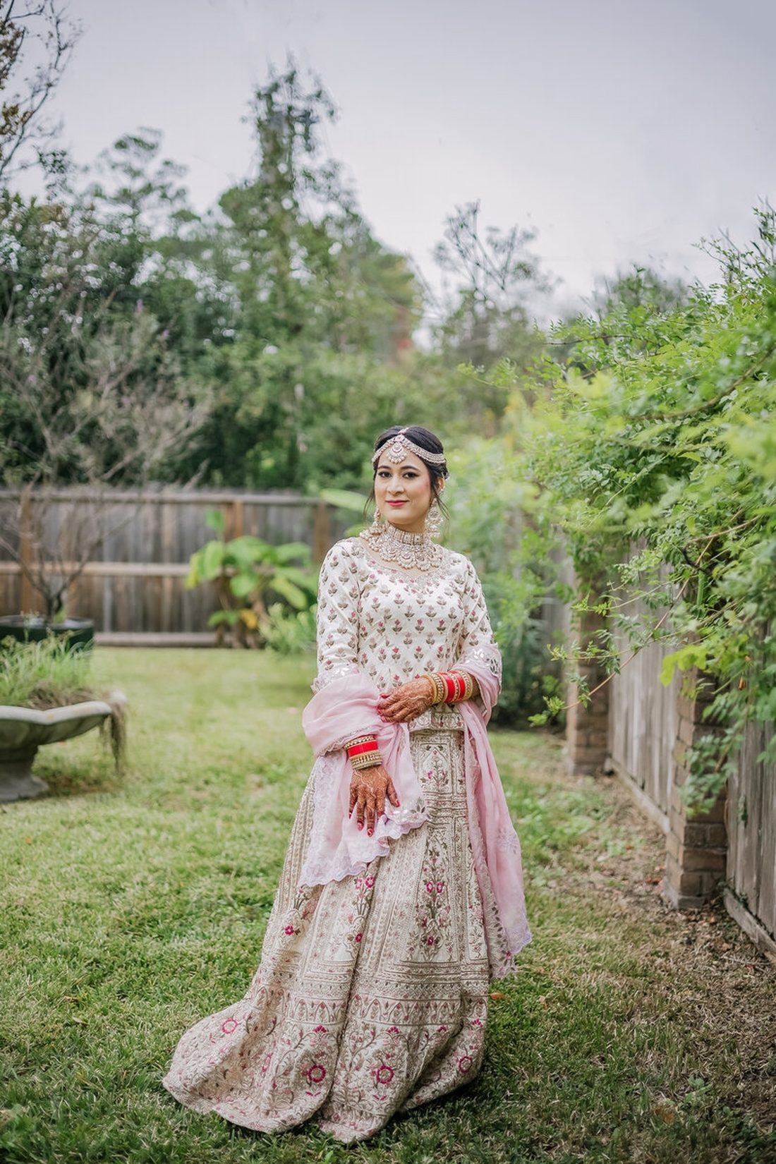 Bride in ivory lehenga and pink dupatta standing among bare-branched garden trees