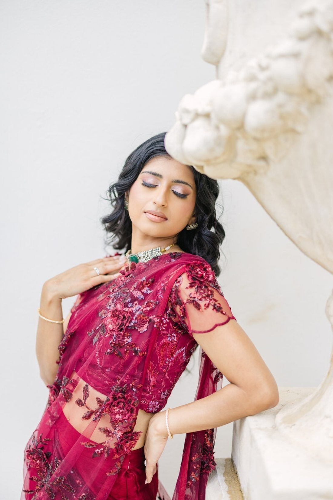 Bride in a deep berry sequined sari leaning against a classical white sculpture