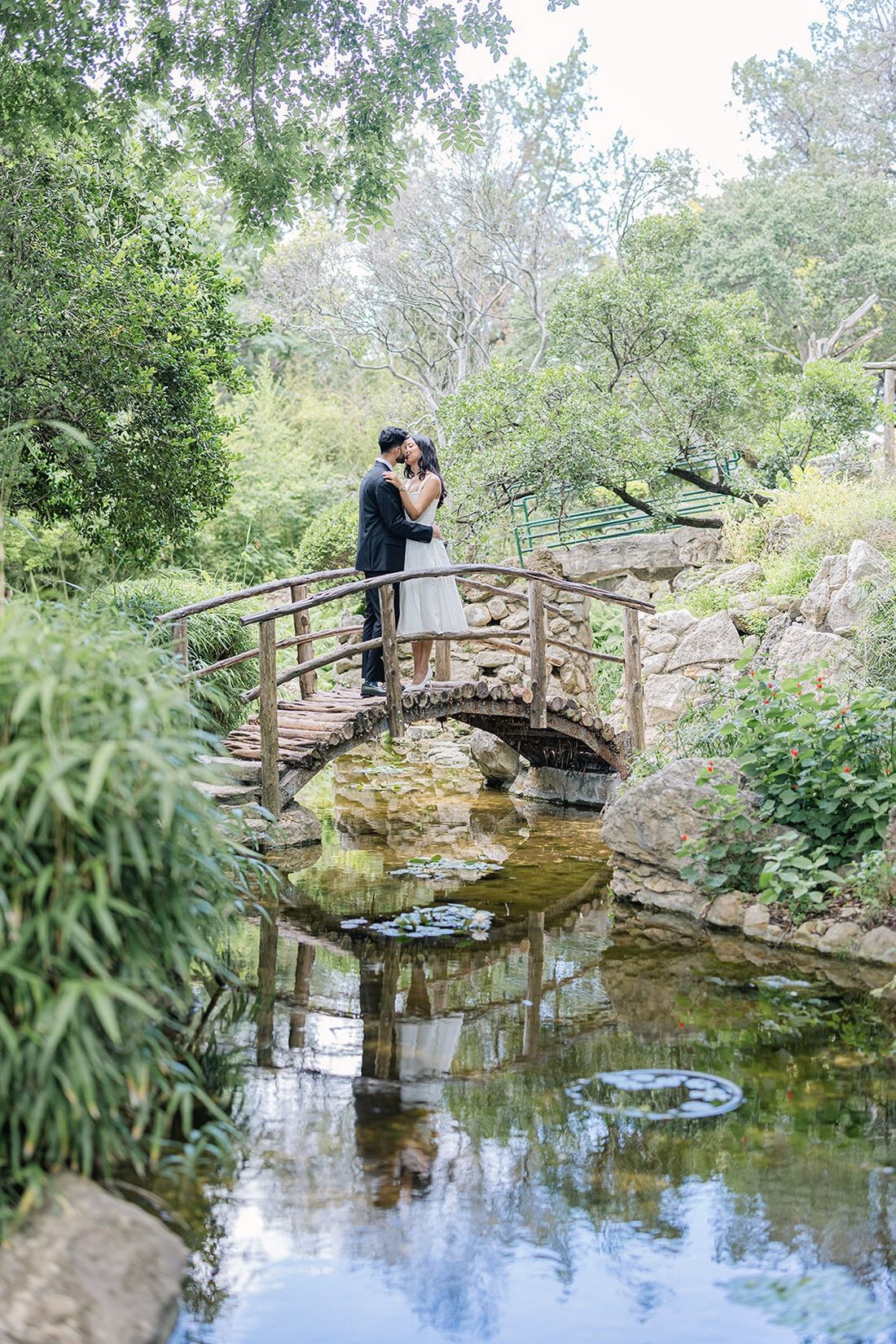 Couple embracing on a small wooden bridge above a koi pond in a lush garden