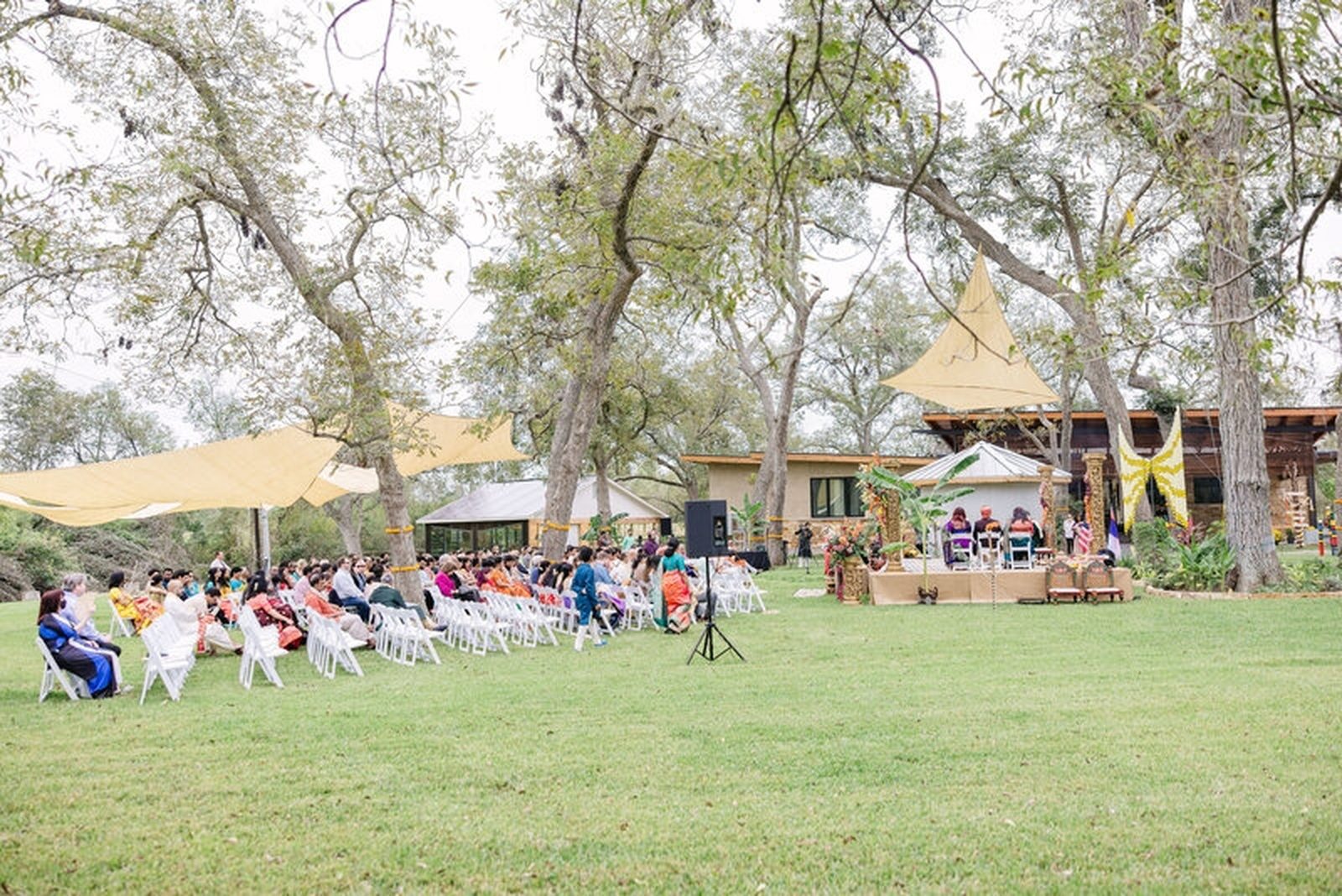 Wide view of an outdoor ceremony with guests seated under shade sails and tall trees
