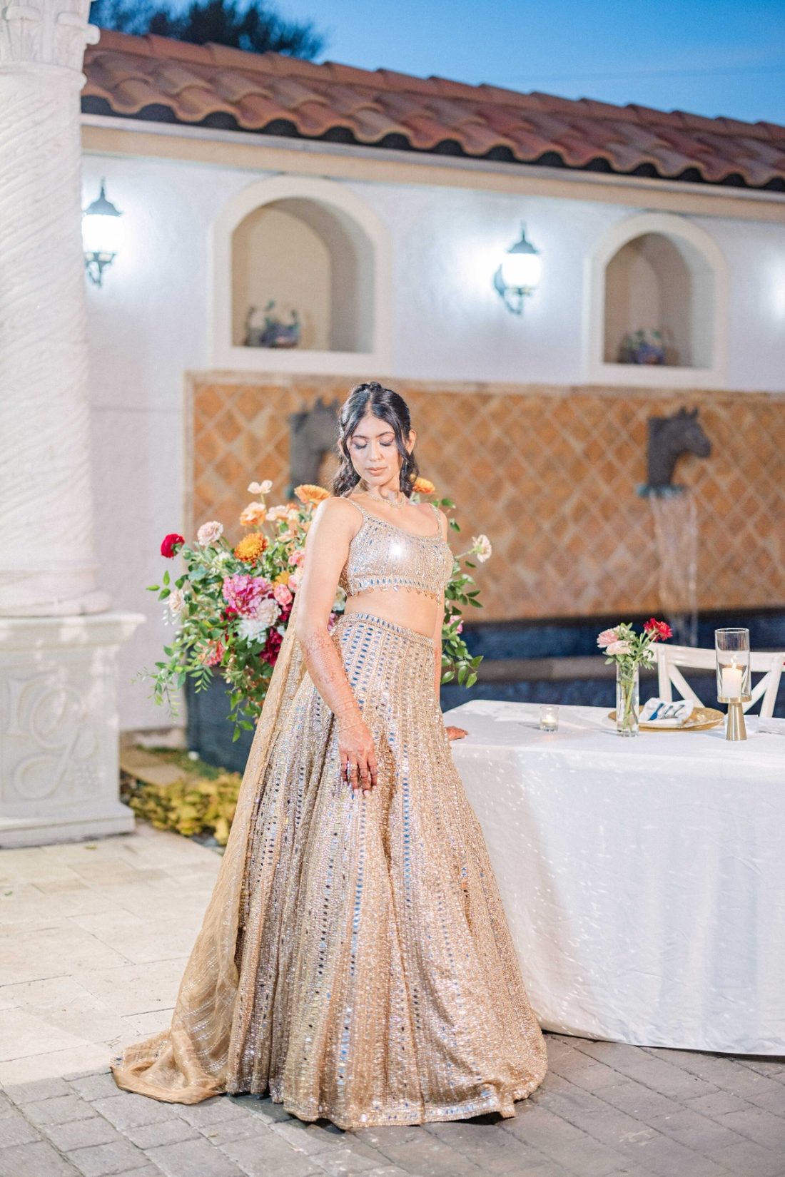 Bride in a shimmering gold reception lehenga in front of a courtyard fountain