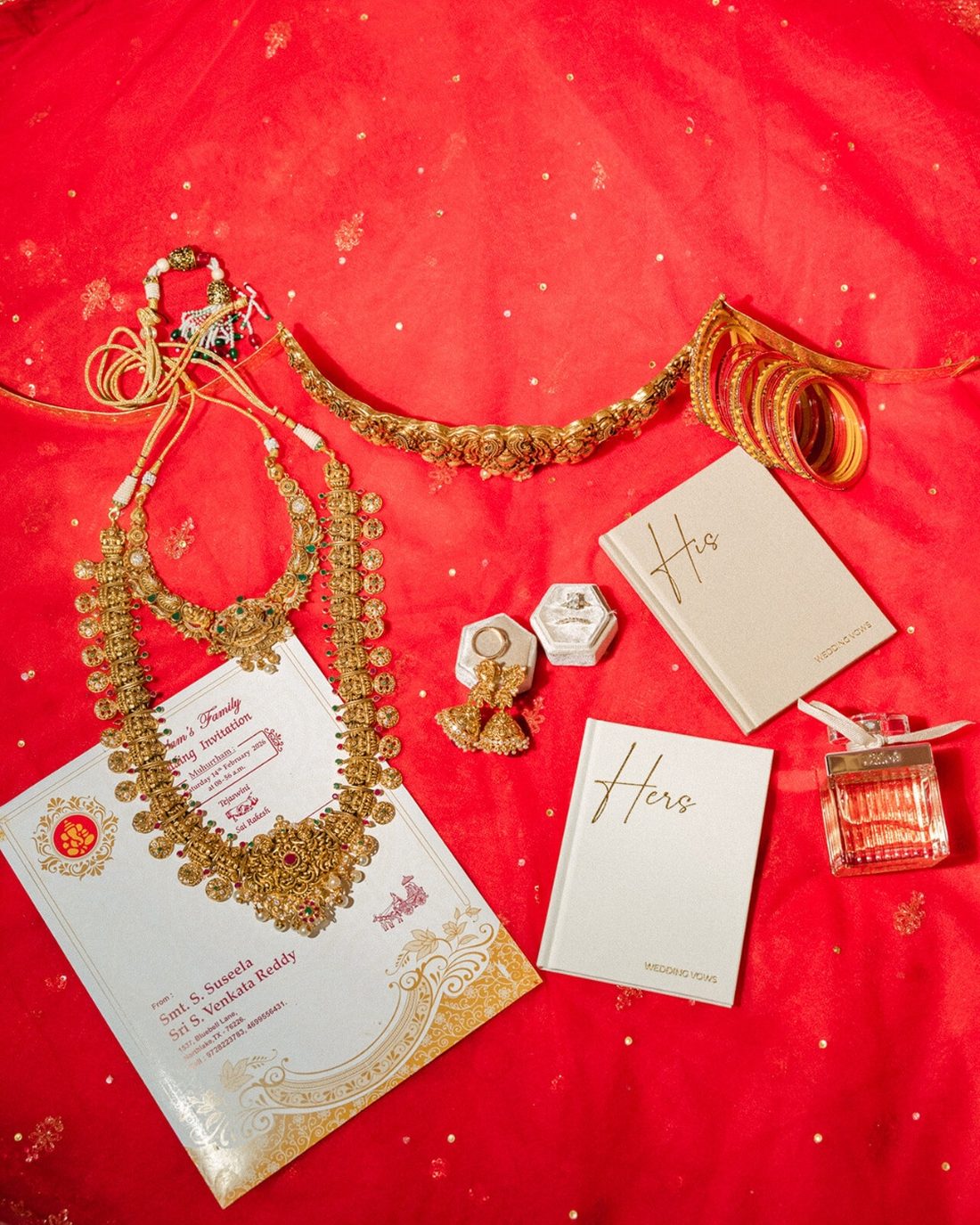 Flat-lay of bridal jewelry, His and Hers vow books, rings, and invitation on a red dupatta